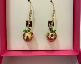 May include: A pair of gold dangle earrings with a red apple charm. The apple has green leaves and a pink rhinestone in the center. The earrings are in a pink box with the text "BETSEY JOHNSON" on the lid.