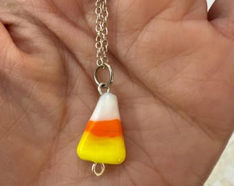 May include: A silver chain necklace with a candy corn pendant. The pendant features the classic candy corn colors: white, orange, and yellow. The necklace is a delicate piece of jewelry.