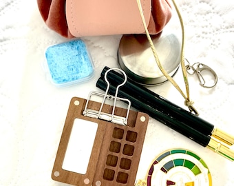 May include: A pink velvet drawstring bag with a gold chain and a metal clasp. The bag is open and there are various items inside, including a wooden palette with a mirror, a small metal container, a blue sponge, two black pens, a gold keychain with a color wheel, and a small notebook with a floral pattern.