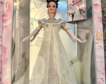 May include: A Barbie doll in a white gown with silver accents, a tiara, and long white gloves, displayed in a box. The box has a silver interior and a pink floral border. A small poster of "My Fair Lady" is visible.