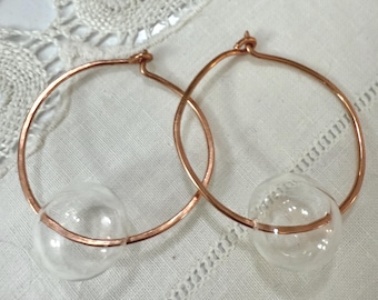 May include: A pair of hoop earrings with a copper-colored metal frame. Each earring features a clear, round glass bead. The earrings are displayed on a white, textured surface.