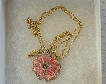 May include: A gold-toned necklace with a pendant shaped like a pink flower with a black center. The flower pendant is hand-painted with a gold outline. The necklace is presented in a white box with a soft, white lining.