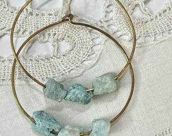 May include: A pair of gold-colored hoop earrings. Each earring features a series of light blue-green, irregularly shaped stones. The earrings are displayed on a white, textured surface.