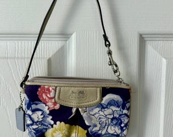 May include: A small Coach wristlet with a floral pattern in blue, yellow, and pink on a navy background. The wristlet has a beige leather accent with the Coach logo and a silver-tone zipper and clasp.