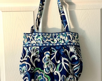 May include: A blue tote bag with a floral pattern in white, green, and light blue. The bag has two handles and a quilted design. The bag is hanging on a silver hook on a white door.