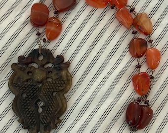 May include: A necklace featuring a carved brown pendant with an intricate design, connected to a chain of orange and red gemstone beads. The beads vary in size and shape, creating a unique and eye-catching accessory.