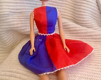 May include: A vintage doll with blonde hair and blue eyes, wearing a red and blue dress with white trim. The dress is divided vertically, with one side red and the other blue. The doll is seated on a textured, light-colored surface.