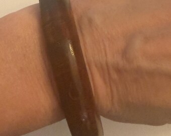 May include: A brown bangle bracelet. The bracelet is a solid, rounded shape with a smooth, glossy finish. The color is a rich, dark brown. The bracelet is worn on a wrist.