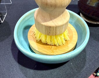 May include: A wooden dish brush with yellow bristles sits in a light blue ceramic bowl. The brush has a round wooden handle and a bamboo base. The bowl is round and has a slightly textured surface. The brush is designed for kitchen use.