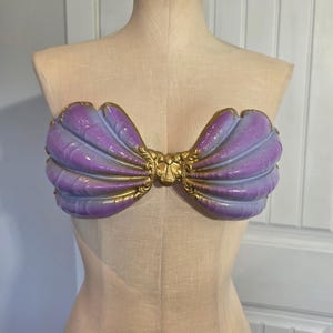 Könnte beinhalten: Ein trägerloses Oberteil in Form von zwei großen Muscheln, das einen Farbverlauf von Violett und Blau mit metallisch goldenem Rand und verzierten goldenen Details in der Mitte aufweist. Die muschelförmigen Cups sind mit Rillen strukturiert.