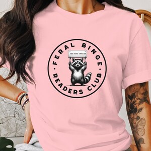 May include: A light pink t-shirt featuring a circular graphic with a raccoon holding a sign that reads "ONE MORE CHAPTER". The graphic is surrounded by the words "FERAL BINGE READERS CLUB" in a black font. The raccoon is black and white.