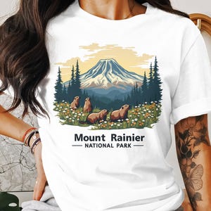 Könnte beinhalten: Weißes T-Shirt mit einem Motiv des Mount Rainier Nationalparks, das eine Bergkette, Bäume und Murmeltiere zeigt. Der Text "Mount Rainier - National Park" ist unter dem Motiv aufgedruckt.