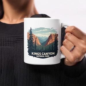 May include: White ceramic mug with a scenic illustration of Kings Canyon National Park. The design features a mountain range, pine trees, and the text "KINGS CANYON NATIONAL PARK". The mug has a curved handle.