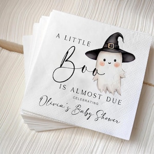 May include: A stack of white paper napkins with a printed design. The design features a cartoon ghost wearing a witch hat, with the text "A Little Boo Is Almost Due Celebrating Olivia's Baby Shower."