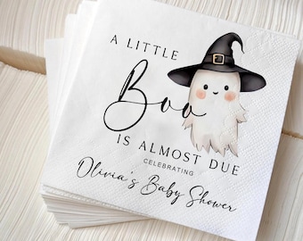 Puede incluir: Una pila de servilletas de papel blancas con un fantasma de dibujos animados que lleva un sombrero de bruja negro. El texto dice A LITTLE Boo IS ALMOST DUE CELEBRATING Olivia's Baby Shower en letras negras con borde texturizado.