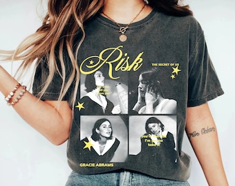 Puede incluir: Una camiseta negra con un estampado gráfico de una mujer cantando en un micrófono. El texto "Risk The Secret of Us" está impreso en la camiseta. El texto "Gracie Abrams" está impreso en la parte inferior de la camiseta.