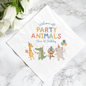 May include: A white square paper napkin featuring the text Welcome all PARTY ANIMALS Olivia 1st Birthday in colorful lettering. The design includes illustrations of a giraffe, crocodile, monkey, and elephant wearing party hats.