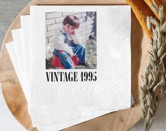 May include: White paper napkins with a diamond pattern and a printed photo of a child in overalls. The photo is above the text "VINTAGE 1995". The napkins are stacked on a wooden tray with a yellow cloth and dried wheat.