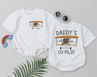 May include: Two white short-sleeved baby onesies. One onesie has a small airplane graphic and the other has a larger airplane graphic with the text "Daddy's Co-Pilot".