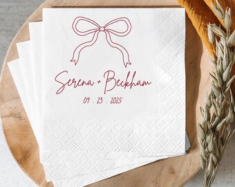 May include: White paper napkins with a burgundy bow design, the names "Serena + Beckham", and the date "09.23.2025" printed on them. The napkins have a diamond pattern and are stacked on a wooden plate.