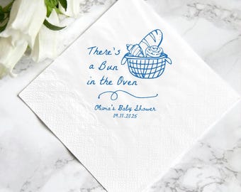 Puede incluir: Servilleta de papel cuadrada blanca con texto azul y una ilustración de una cesta de pan. El texto dice "There's a Bun in the Oven" y "Olivia's Baby Shower 09.11.2025". La servilleta está sobre una superficie de mármol.