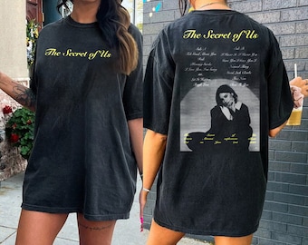 May include: Black oversized t-shirt with the text "The Secret of Us" in yellow on the front and a black and white photo of a woman with the same text in yellow on the back.
