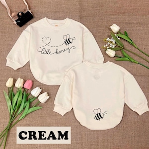 May include: Cream-colored baby sweatshirt with long sleeves. The front features a black and white bee graphic with the words "little honey." The back of the sweatshirt has a bee graphic. The word "CREAM" is at the bottom of the image.