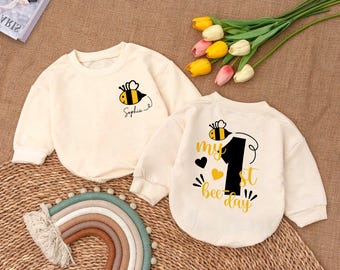 Op de afbeelding: Twee crèmekleurige shirts met lange mouwen met bijen- en nummer één-ontwerpen. Het eerste shirt heeft een bij met de tekst "Sophia" eronder. Het tweede shirt heeft een bij met de tekst "my 1st bee day".