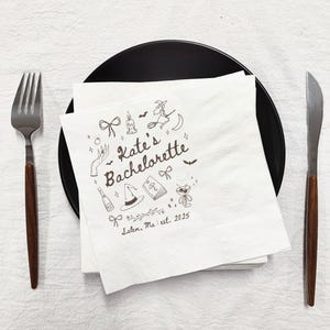 May include: A white paper napkin with a black plate, fork, and knife. The napkin has a black illustration with the text "Kate's Bachelorette" and the location "Salem, Ma. est. 2025". The illustration includes a witch on a broom, a witch hat, and other festive elements.