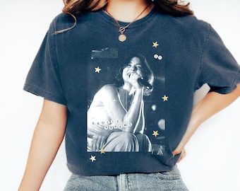 May include: A dark blue t-shirt with a black and white photo of a woman singing on stage. The text "GRACIE ABRAMS" is printed in white letters on the shirt. There are gold stars printed on the shirt.