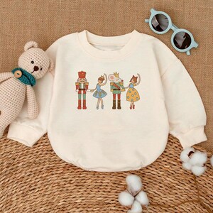 May include: A cream-colored sweatshirt with a vintage-style illustration of a nutcracker, a mouse king, and two ballerinas. The illustration is in shades of red, blue, green, and yellow.