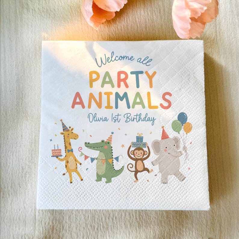 May include: A white square paper napkin featuring the text Welcome all PARTY ANIMALS Olivia 1st Birthday in colorful letters. Below the text are illustrations of a giraffe, crocodile, monkey, and elephant wearing party hats and holding gifts.