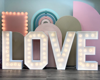 Può includere: Lettere bianche illuminate che compongono la parola "LOVE" con lampadine in ogni lettera. Le lettere sono davanti a uno sfondo rosa, beige e blu.