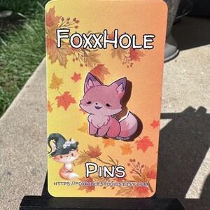 May include: A rectangular pin featuring a cartoon fox with the text "FoxxHole" and "Pins". The background is orange with autumn leaves. A small fox wearing a witch hat is also featured. The Etsy shop URL is visible at the bottom.