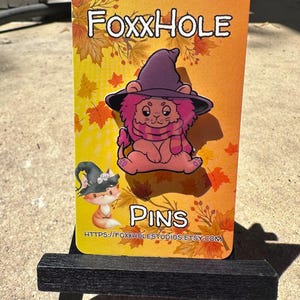 Op de afbeelding: Een roze leeuw met een paarse heksenhoed en een sjaal, op een witte achtergrond met herfstbladeren en de tekst "Foxxhole Pins" en een websiteadres.