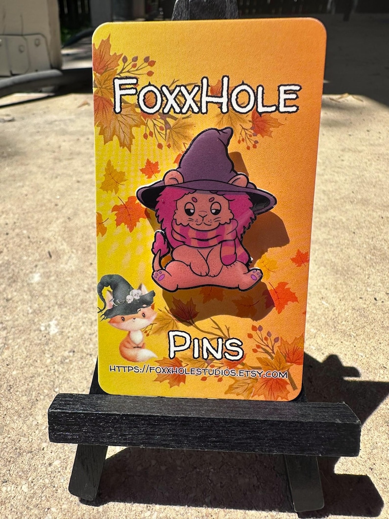 Op de afbeelding: Een roze leeuw met een paarse heksenhoed en een sjaal, op een witte achtergrond met herfstbladeren en de tekst "Foxxhole Pins" en een websiteadres.
