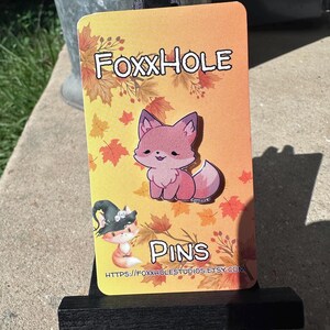 May include: A rectangular card with the text "FoxxHole Pins" and a cartoon fox illustration. The card has a gradient orange background with autumn leaves. A small black easel supports the card.