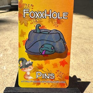 Op de afbeelding: Een gele kaart met de tekst "FOXXHOLE PINS" en een websiteadres. De kaart toont een cartoonillustratie van een blauwe slang die zich oprolt met een paarse tong.