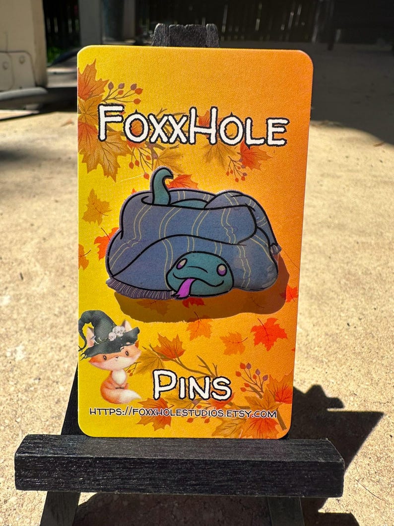 Op de afbeelding: Een gele kaart met de tekst "FOXXHOLE PINS" en een websiteadres. De kaart toont een cartoonillustratie van een blauwe slang die zich oprolt met een paarse tong.