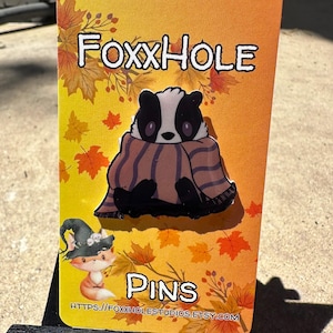 Op de afbeelding: Een kleurrijke kaart met de tekst "Foxxhole Pins" en een cartoon-das die een gestreepte sjaal draagt. De achtergrond heeft een herfstthema met oranje, gele en rode bladeren.
