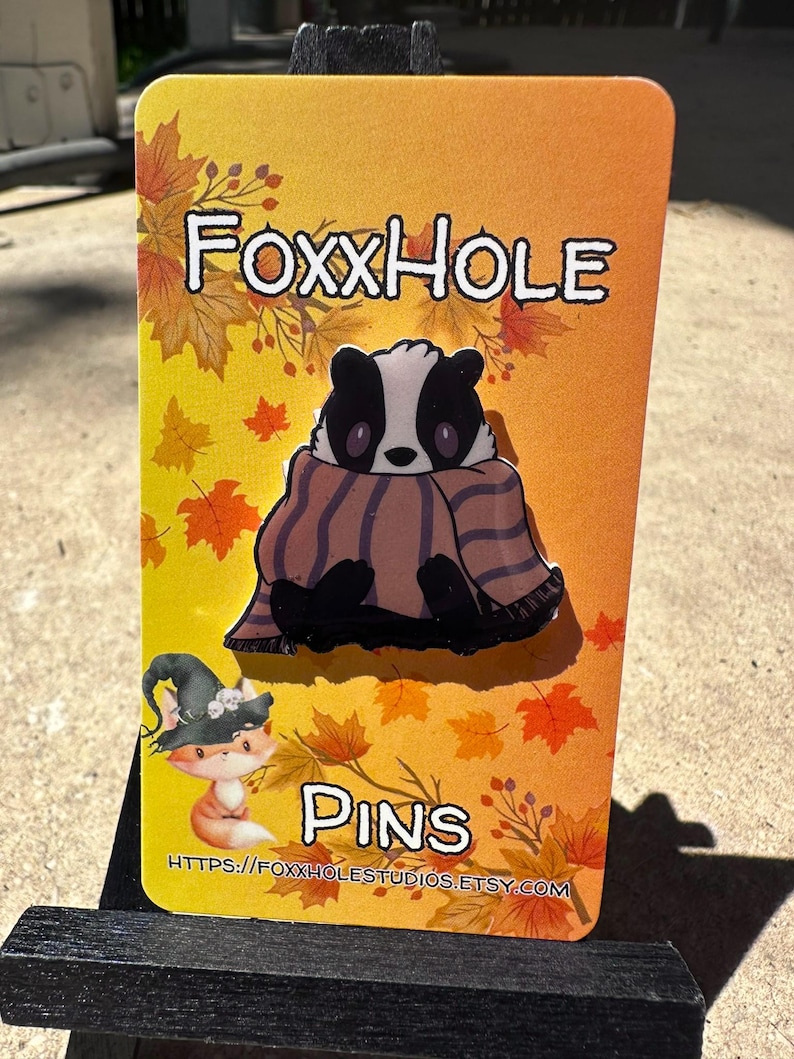 Op de afbeelding: Een kleurrijke kaart met de tekst "Foxxhole Pins" en een cartoon-das die een gestreepte sjaal draagt. De achtergrond heeft een herfstthema met oranje, gele en rode bladeren.