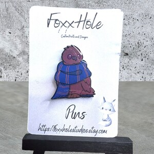 Op de afbeelding: Een bruine en paarse emaille pin van een uil met een blauw-rode gestreepte sjaal. De pin is op een witte kaart met de tekst "FoxxHole Custom Prints and Designs" en "Pins" en het websiteadres "https://foxxholestudios.etsy.com".