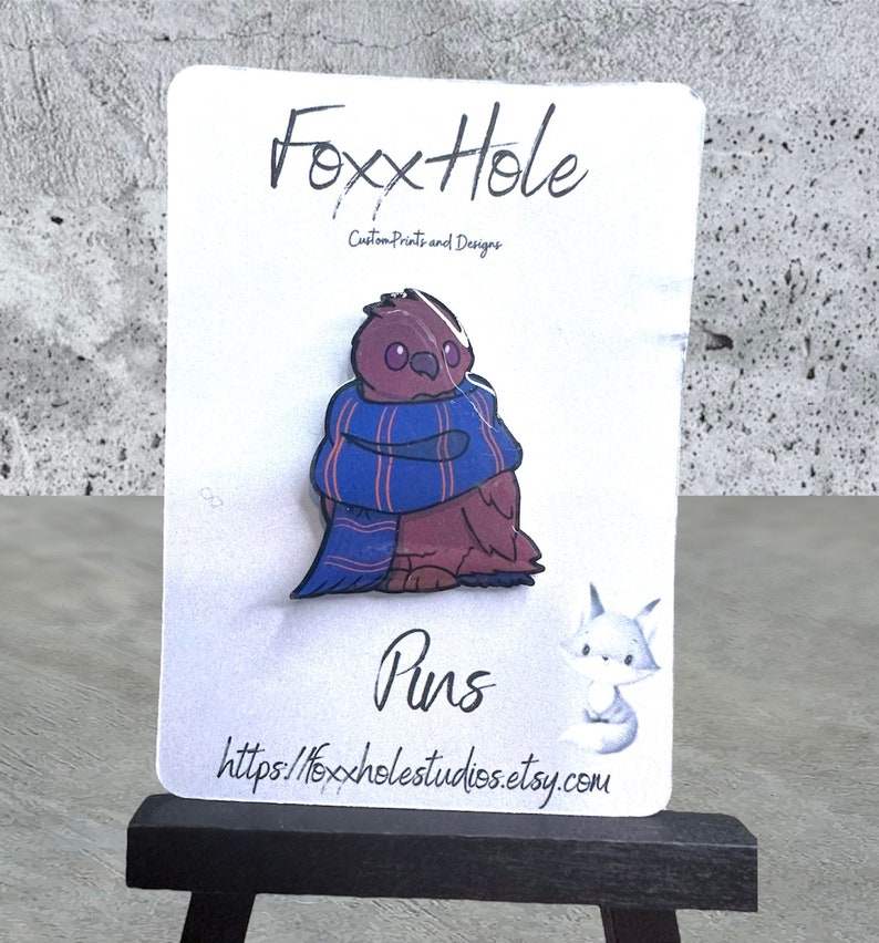 Op de afbeelding: Een bruine en paarse emaille pin van een uil met een blauw-rode gestreepte sjaal. De pin is op een witte kaart met de tekst "FoxxHole Custom Prints and Designs" en "Pins" en het websiteadres "https://foxxholestudios.etsy.com".