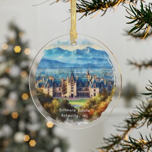 May include: A round glass ornament with a watercolor image of Biltmore Estate in Asheville, North Carolina. The image shows the large house with a mountain range in the background.