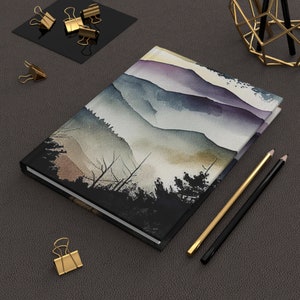 May include: A black hardcover journal with a watercolor painting of a mountain range with purple, blue, and green hues. The mountains are in the background and there are trees in the foreground.