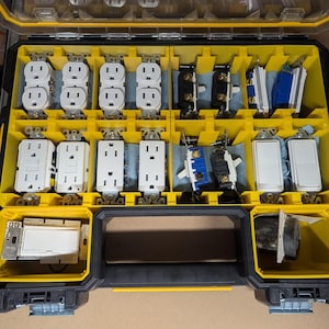 Może przedstawiać: Żółto-czarna plastikowa skrzynka narzędziowa z różnymi elementami elektrycznymi, w tym włącznikami światła i gniazdami elektrycznymi, uporządkowanymi w przegródkach.