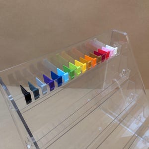 May include: A clear acrylic tiered display stand featuring a row of small, rectangular, multi-coloured plastic clips arranged in a spectrum. The stand has multiple slanted shelves designed for organisation, with the colourful clips lined up neatly.
