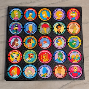 May include: A collection of 25 circular collectible cards featuring characters from The Simpsons animated television series. Each card has a colorful illustration of a character's face and name. The cards are arranged in a black display case.