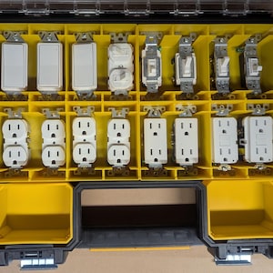 May include: A yellow and black toolbox filled with various electrical switches and outlets. The switches are white with silver metal components. The outlets are white with black slots. The toolbox is divided into compartments.