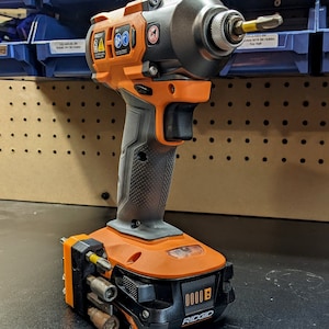 Puede incluir: Un destornillador de impacto inal&aacute;mbrico naranja y negro con un soporte de brocas integrado. El destornillador tiene una bater&iacute;a negra con un logotipo RIDGID amarillo y un indicador de nivel de bater&iacute;a. El destornillador est&aacute; sobre una superficie negra.